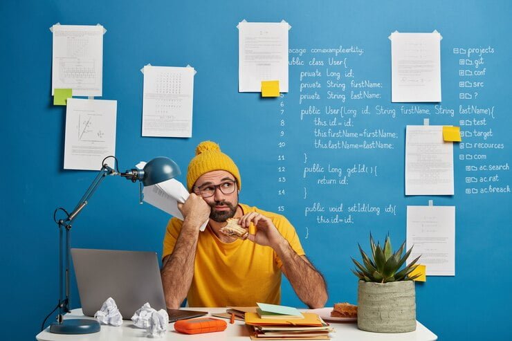 thoughtful-male-programmer-software-developer-ponders-program-code-looks-away-eats-burger-holds-papers-wears-yellow-clothes-spends-time-making-projec